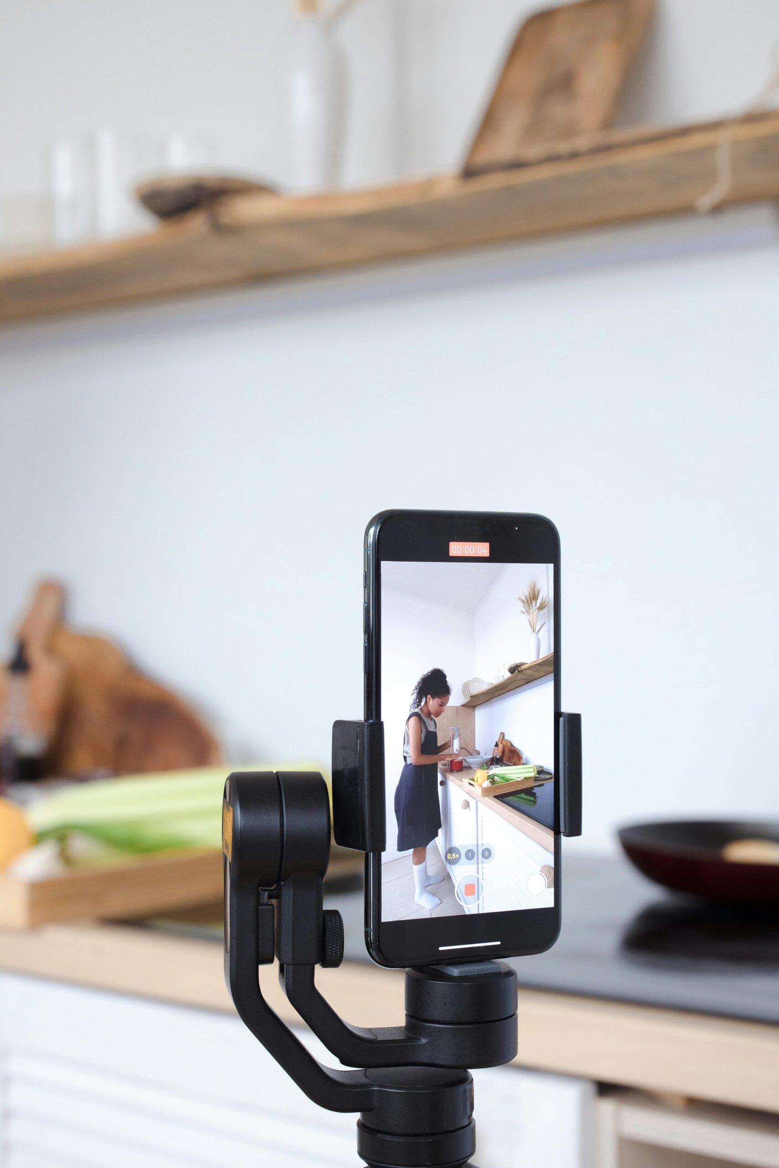 A content creator uses a smartphone and tripod to film a cooking session in a modern kitchen setting.
